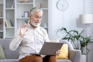 Cheerful elderly man having a chat using a laptop from the comfort of his living room, waving hello. photo