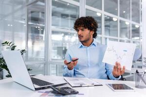 Upset financier showing online report, man working inside office nervously showing document, hispanic man in online meeting using laptop for remote communication and call. photo