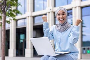emocionado musulmán mujer en hijab se sienta al aire libre con su computadora portátil, celebrando un profesional éxito con un jubiloso expresión cerca un oficina edificio. foto