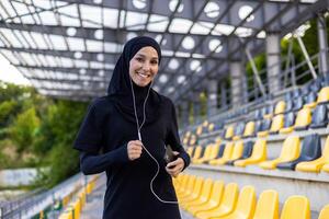 positivo islámico hembra en hijab y juguetón ropa haciendo ejercicios con cableado auriculares conectado a móvil teléfono. sonriente dama disfrutando gasto hora a estadio y practicando corriendo a música. foto