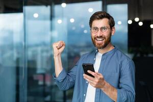 retrato de un moderno empresario en el medio de el oficina, el hombre es mirando a el cámara con un teléfono inteligente y celebrando el victoria, el hombre es participación el teléfono en su manos y levantamiento su mano en un gesto de triunfo. foto