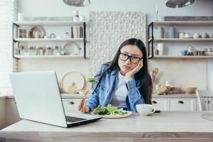 Dissatisfied and tired of dieting young Asian woman. He is sitting at the table in the kitchen, watching a nutritionist's webinar on a laptop. He holds a fresh salad, looks into the camera. photo