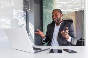 call online meeting mature african american businessman talking to colleagues remotely using laptop, man in business suit smiling gesturing cheerfully with hands inside office. photo