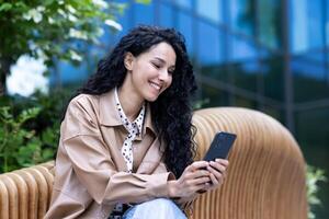 alegre hermosa mujer sentado en banco fuera de oficina edificio, exitoso latín americano negocio mujer utilizando teléfono, leyendo Noticias en línea y hojeada en línea páginas, participación teléfono inteligente foto