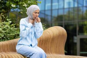 Muslim woman coughing and feeling sick while having pulmonary disease and sitting outdoors. Struggling lady covering mouth with clenched fist for not spreading virus around in downtown district. photo