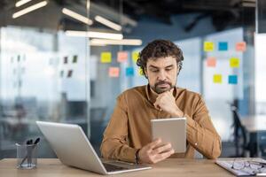 A contemplative man in casual business attire working on a laptop while holding a tablet in a modern office setting, surrounded by colorful sticky notes. photo