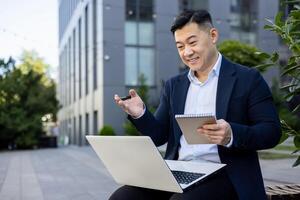 Smiling young Asian male businessman in a suit sitting outside an office building, holding a laptop on his lap, talking on a call and making notes in a notebook. photo