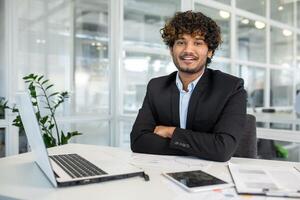 Confident young male entrepreneur with curly hair smiling in a well-lit office. Laptop and documents on table, reflecting a productive business environment. photo