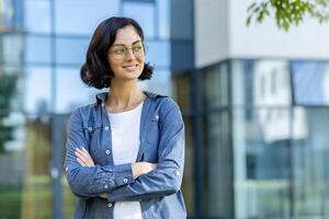 un joven hembra estudiante con un confidente sonrisa, en pie brazos cruzado en frente de un moderno educativo edificio, vistiendo casual atuendo. foto