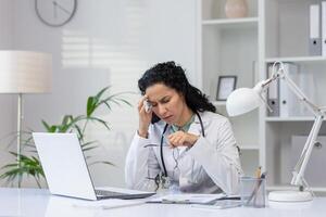 A worried Hispanic female doctor appears stressed while talking on the phone in her office, exuding an air of concern and concentration. photo