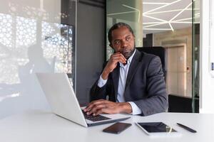 Thinking mature adult african american businessman at workplace, financier boss investor with laptop thinking about financial solutions new projects inside office. photo