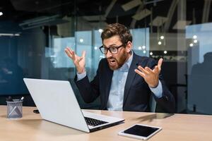 Angry and angry businessman talking on call, talking to colleagues and shouting quarrel, mature boss working inside modern office building, senior man with laptop and beard. photo
