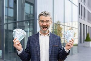 Businessman is delighted with a huge win, holds stacks of dollars and shouts with joy, looks at the camera. photo