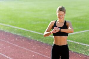 hembra atleta sonriente y comprobación resultados de aptitud formación y corriendo en reloj inteligente pulsera, mujer atleta sonriente comprobación corazón Velocidad en dispositivo mientras corriendo en estadio en soleado día. foto