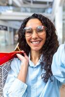 hermosa Hispano mujer mirando dentro teléfono inteligente cámara hablando en llamada y tomando selfie foto, compras en grande Tienda participación vistoso regalo pantalones en manos sonriente hablando con amigos foto