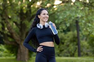 sonriente activo mujer en rutina de ejercicio engranaje tomando un descanso con un agua botella en un verde parque configuración, auriculares alrededor su cuello. foto