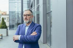 retrato de grave mayor gris peludo hermoso exitoso empresario en lentes y traje. él soportes cerca el oficina centro, doblada su brazos en frente, mira a el cámara, sonrisas foto