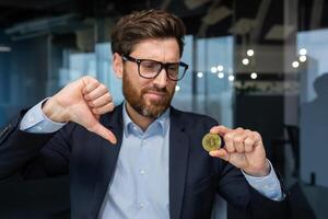 Close-up photo of male businessman holding gold crypto currency bitcoin coin in hands, mature investor disappointed with money rate and investment, boss in business suit showing finger down.