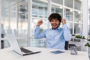 alegre joven empresario con Rizado pelo emocionado celebrando un victoria o éxito mientras trabajando en su ordenador portátil en un brillante, contemporáneo oficina espacio. foto