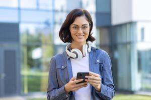 A stylish young woman in glasses and casual attire enjoys using her smartphone, with headphones around her neck, standing outdoors with a modern building background. photo