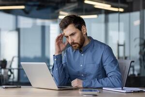 A serious and thoughtful young man sits in the office at the desk, leans his head on his hand and looks at the screen of the laptop monitor, solving a question and a problem. photo