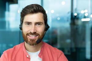 Close-up portrait of mature man with beard and call headset inside office, tech support worker smiling and looking at camera in red shirt, online customer service and support. photo