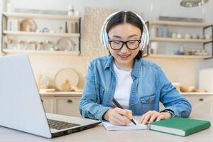 Online training in quarantine. Young beautiful Asian student girl sitting at the table at home wearing white headphones and using a laptop. He studies remotely. Writes in a notebook. photo