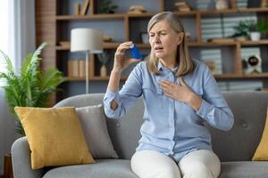 Grey haired woman in light outfit holding arm on chest while using medication for respiratory problem in living room. Disturbed patient experiencing asthma attack and relieving breathing struggles. photo