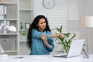 profesional mujer a trabajo haciendo muecas en dolor mientras agarrando su codo, posiblemente sufrimiento desde un lesión o crónico condición. foto