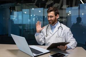 Mature doctor working remotely from modern clinic office, man inside talking on call with patient using laptop photo