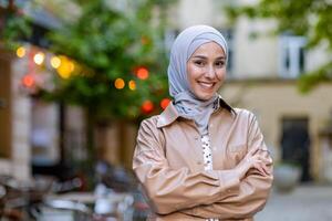 elegante islámico hembra en beige chaqueta en pie con brazos doblada en cofre en borroso antecedentes de patio. retrato de mujer en hijab sensación confidente y mirando a cámara con con dientes sonrisa. foto