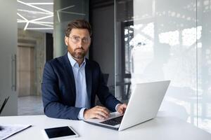 Portrait of a serious young businessman sitting in the office at a desk, working on a laptop and with documents, looking confidently at the camera. photo