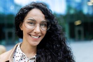 Portrait of a young Hispanic businesswoman with curly hair and glasses smiling during a work break outside an urban office building. photo