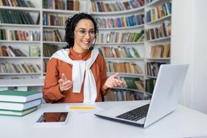 Teacher conducts lessons online, woman with call headset smiling and explaining lecture online using laptop for remote communication and learning, sitting at desk in university library at desk. photo