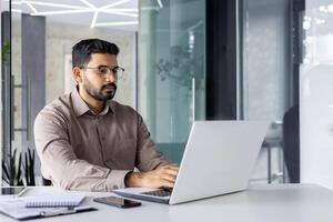 Focused Indian man in smart casual attire working and thinking seriously while using a laptop in a modern office setup. photo