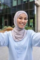 un moderno mujer en un hijab capturas un selfie con un fondo de urbano arquitectura, encarnando confianza y urbano estilo de vida. foto