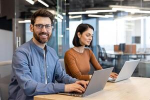 Positive male in glasses looking at camera with toothy smile while sitting by pc next to brunette woman with short haircut. Optimistic trainee enjoying working process with help of experienced mentor. photo