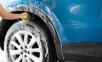 A person is washing a car tire with a green brush. Tire is covered in water and soap. Blue car wash with white soap foam. Auto care service. Car cleaning service concept. Vehicle cleaning service. photo