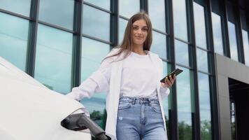 giovane donna è in piedi vicino il elettrico auto e sembra a il inteligente Telefono. il noleggio auto è ricarica a il ricarica stazione per elettrico veicoli. auto condivisione video