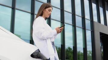 joven mujer es en pie cerca el eléctrico coche y mira a el inteligente teléfono. el alquiler coche es cargando a el cargando estación para eléctrico vehículos coche compartiendo video