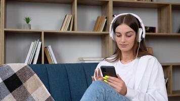 cabeça tiro sorridente pacífico mulher relaxante em confortável sofá, ouvindo para favorito audio música dentro fones de ouvido. jovem feliz menina desfrutando salão estresse livre Tempo às casa video