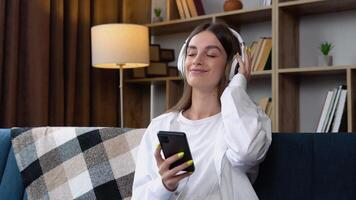 Head shot smiling peaceful woman relaxing on comfortable couch, listening to favorite audio music in headphones. Young happy girl enjoying lounge stress free time at home video