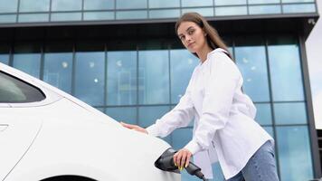 Smiling young caucasian girl plugging electricity cable in electric vehicle for charging on sunny mall parking. Lifestyle and ecology concept video