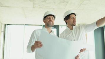 A young engineers in safety helmets standing on an under-construction building holding a blueprint of the structure and working together video
