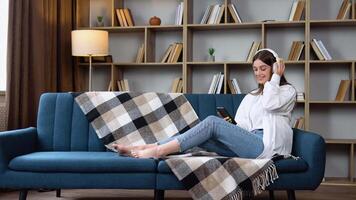 Head shot smiling peaceful woman relaxing on comfortable couch, listening to favorite audio music in headphones. Young happy girl enjoying lounge stress free time at home video