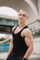 A man with a muscular build stands in front of a bridge photo