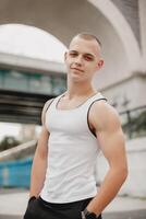 A man with a bald head and a white tank top is standing in front of a bridge photo