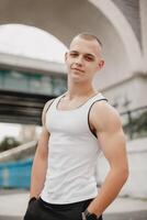 un hombre con un calvo cabeza y un blanco tanque parte superior es en pie en frente de un puente foto