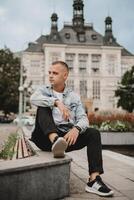 A man in a denim jacket sits on a bench in front of a large building photo