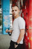 A man in a white shirt and black pants stands in front of a graffiti wall photo